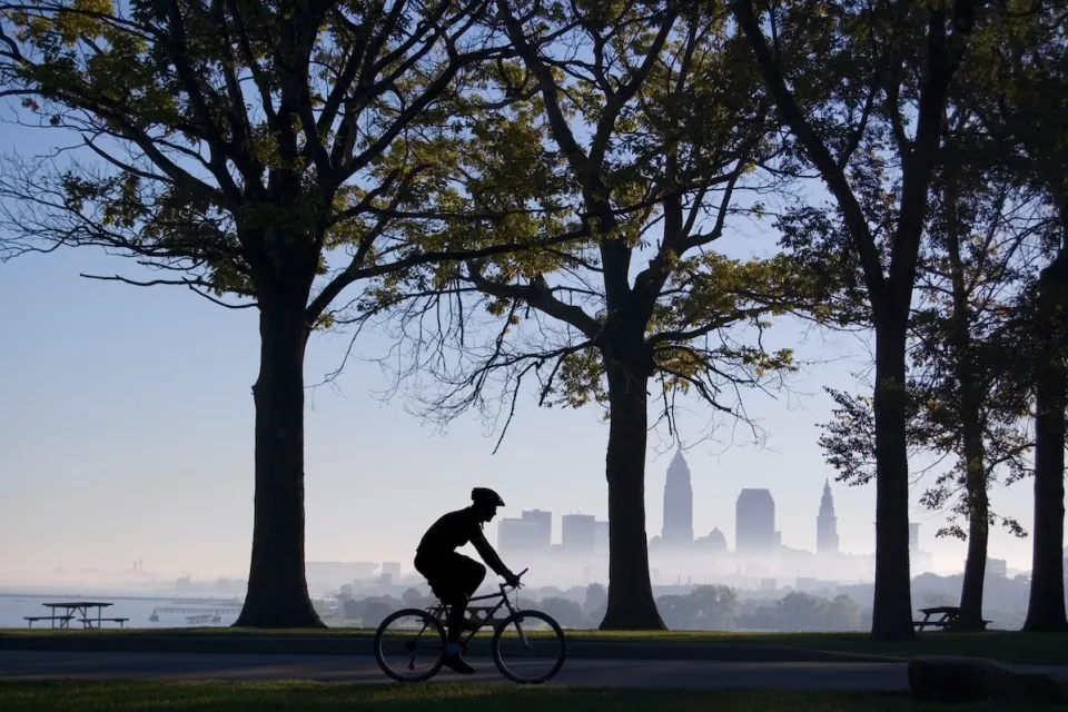 Sillhouette of cyclist riding on path among trees with downtown Cleveland, Ohio skyline visible across water in background.